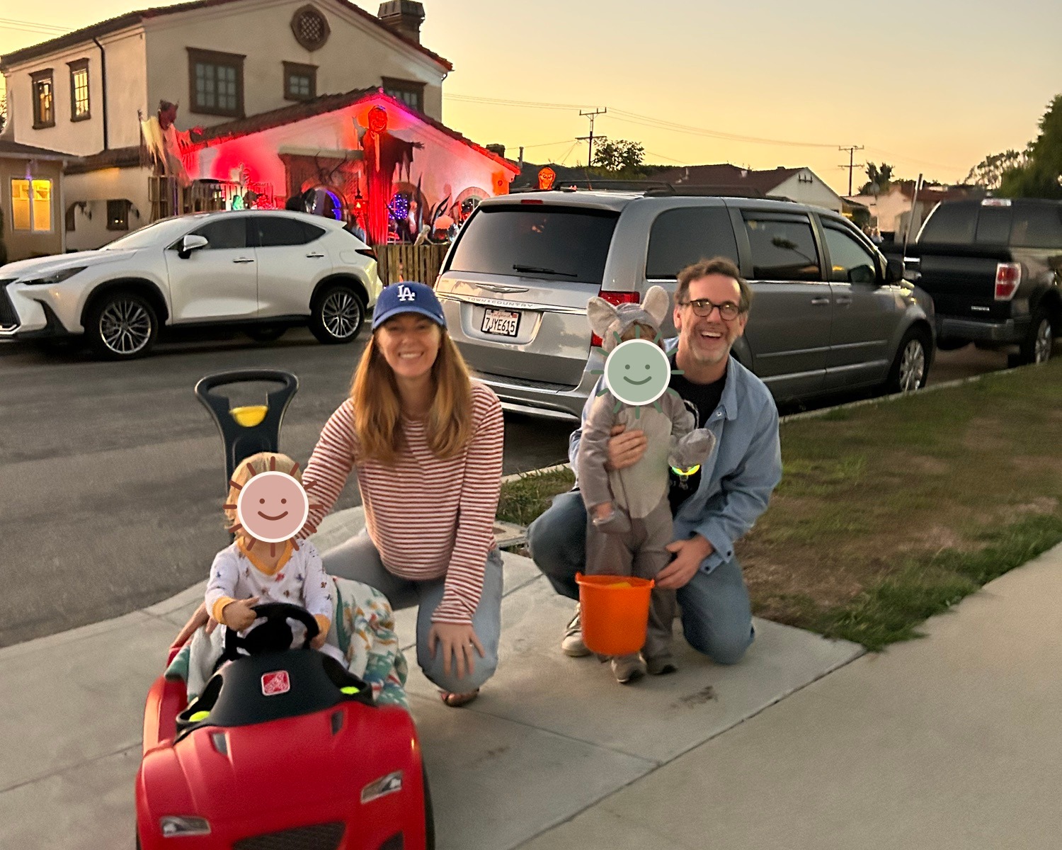The family trick-or-treating together in front of a decorated house, with the children's faces covered by illustrated stickers for privacy.